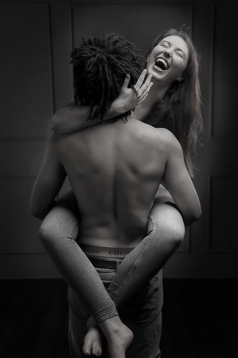 Schwarzweiss Aktfoto eines glücklichen jungen Paares Sie umklammert Ihn mit Ihren Beinen und lacht herzlich.