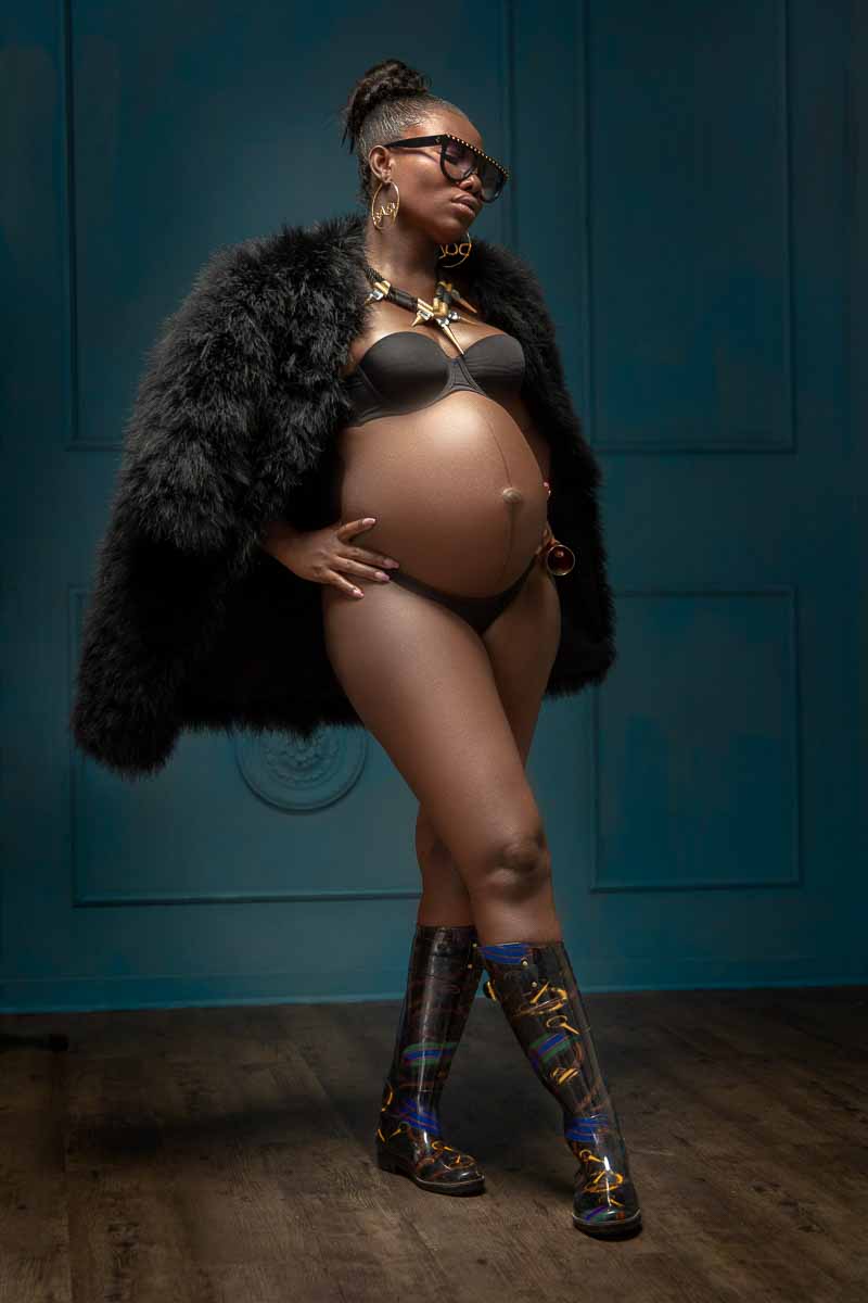 Fotoshooting einer schwangeren dunkelhäutigen Frau, Gangster Ghetto-Outfit steht in Gummistiefeln vor einer blauen klassischen Wand