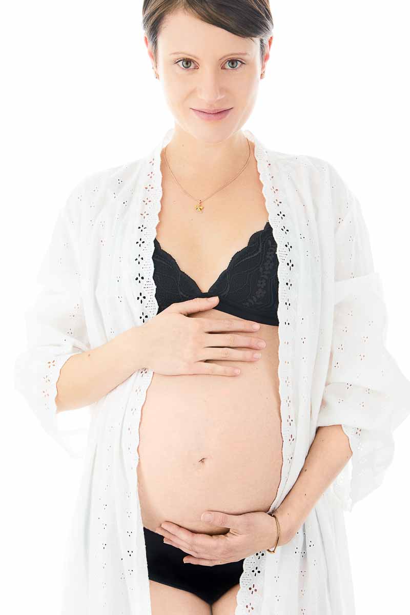 Fotoshooting einer schwangeren kurzhaarigen jungen Frau, die glücklich unter einer weißen romantischen Bluse ihren Babybauch zeigt
