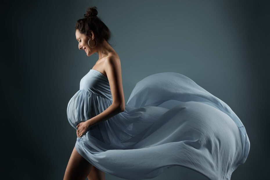 Fotoshooting einer schwangeren, glücklich lachenden, schwarzhaarigen Frau in einem türkis-blauem Kleid, das elegnat weht.
