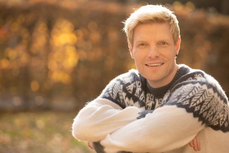 Dating-Foto eines lächelnden jungen blonden Mannes in einem warmen, hellen Strickpullover vor herbstlichem Hintergrund