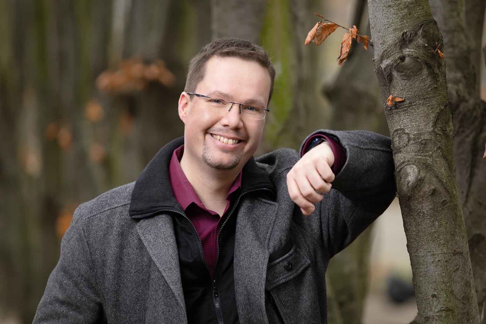 Dating-Foto eines jungen Mannes in einer warmen Jacke, der sich freundlich lächelnd an einem Baum lehnt