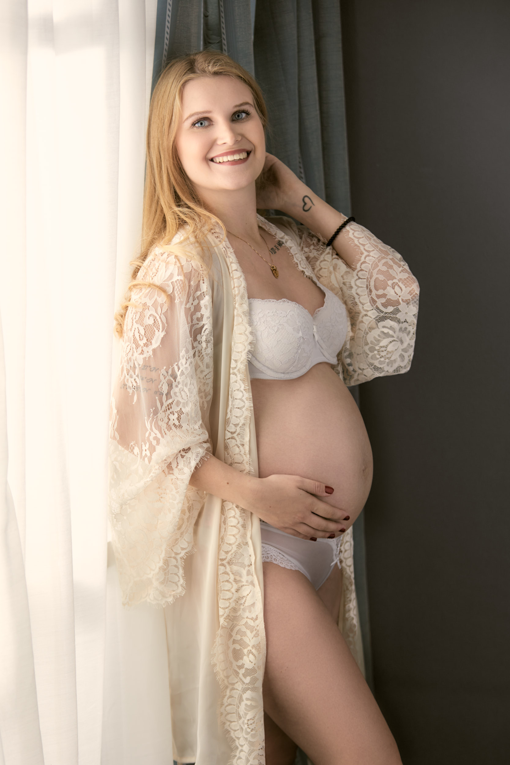 Fotoshooting einer schwangeren, glücklich lachenden, blonden jungen werden Mutter mit Babybauch in einem champagnerfarbenem Spitzenmantel, die an einem hellen Fenster lehnt.