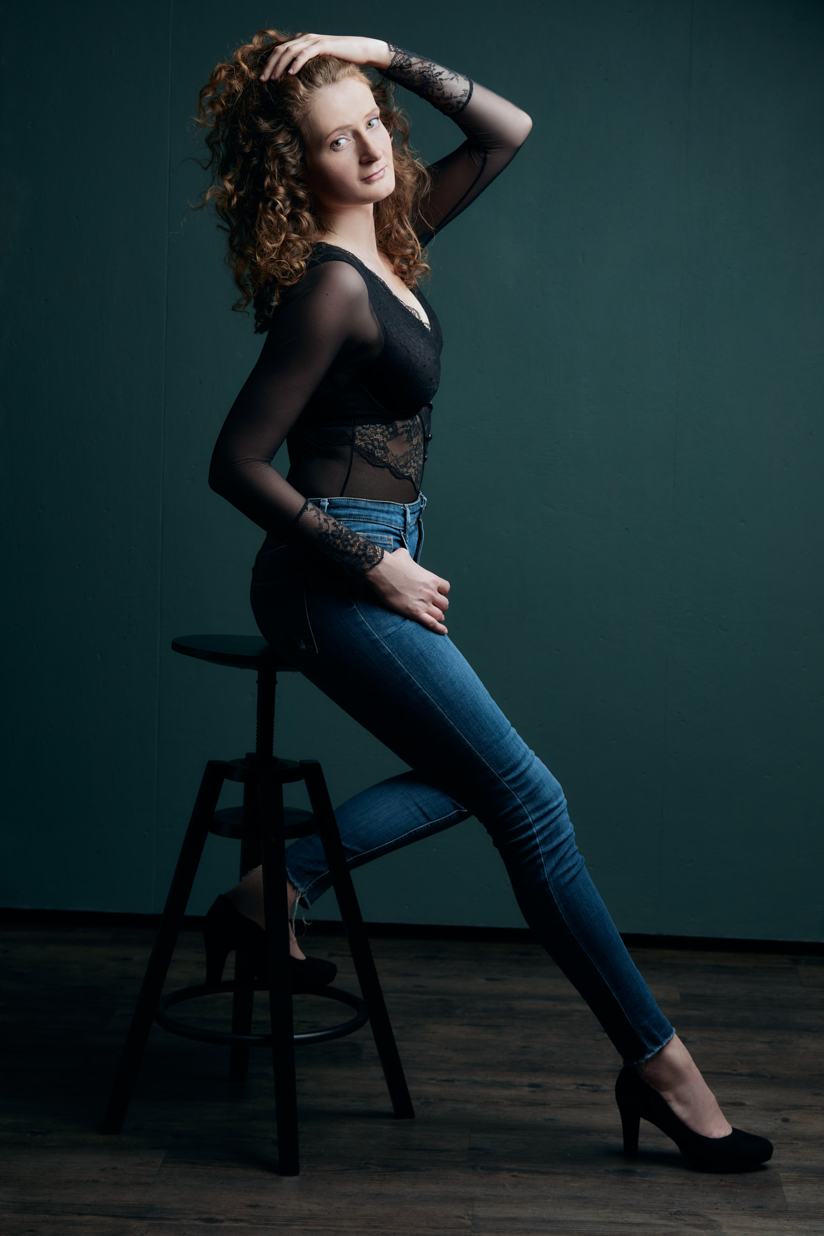Akt-Fotoshooting einer lockigen Frau die in schwarzer Body und blauer Jeans vor einer petrolfarbenen Wand auf einem Barhocker sitz und sich durch die Haare fährt.