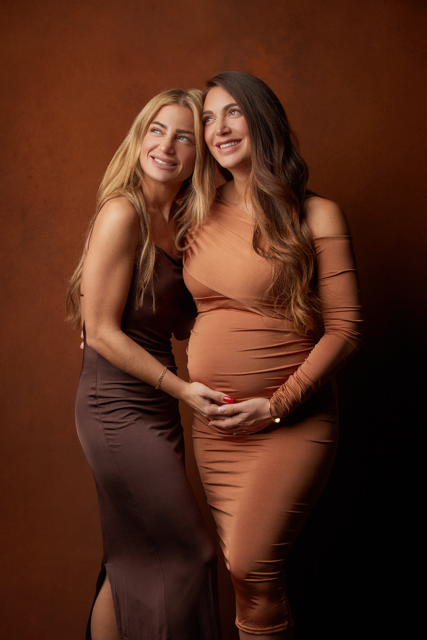 Schwangerschaftsfotoshooting einer schwangeren, glücklich lachenden, braunhaarigen jungen werden Mutter mit Babybauch die gemiensam mit Ihrer blonden Freundin den Bauch hält