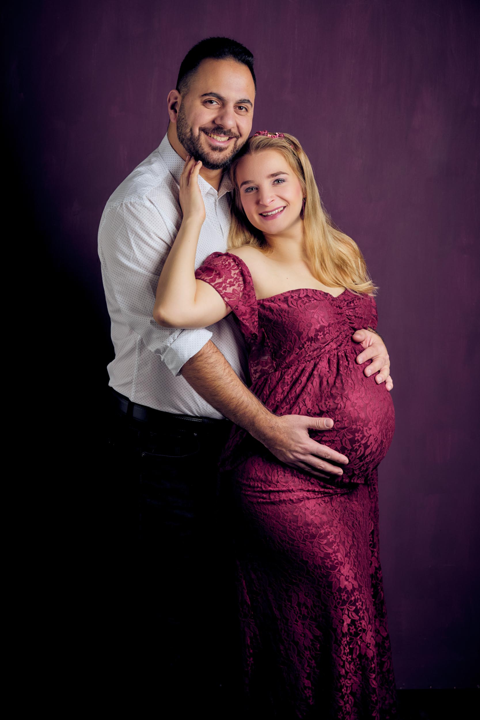 Schwangerschaftsfotoshooting einer schwangeren, glücklich lächenden, bloden jungen werden Mutter mit Babybauch in einem violetten Spitzenkleid wird liebevoll von dem werdenden Vater im weißen Hemd im Arm gehalten.