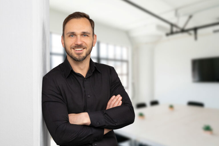 Modernes Bewerbungsfoto eines freundlich lachenden Mannes mit Dreitagebart und schwarzem Hemd der mit verschränkten Armen in einem hellen Sitzumgsraum an einer weißen Wand lehnt.