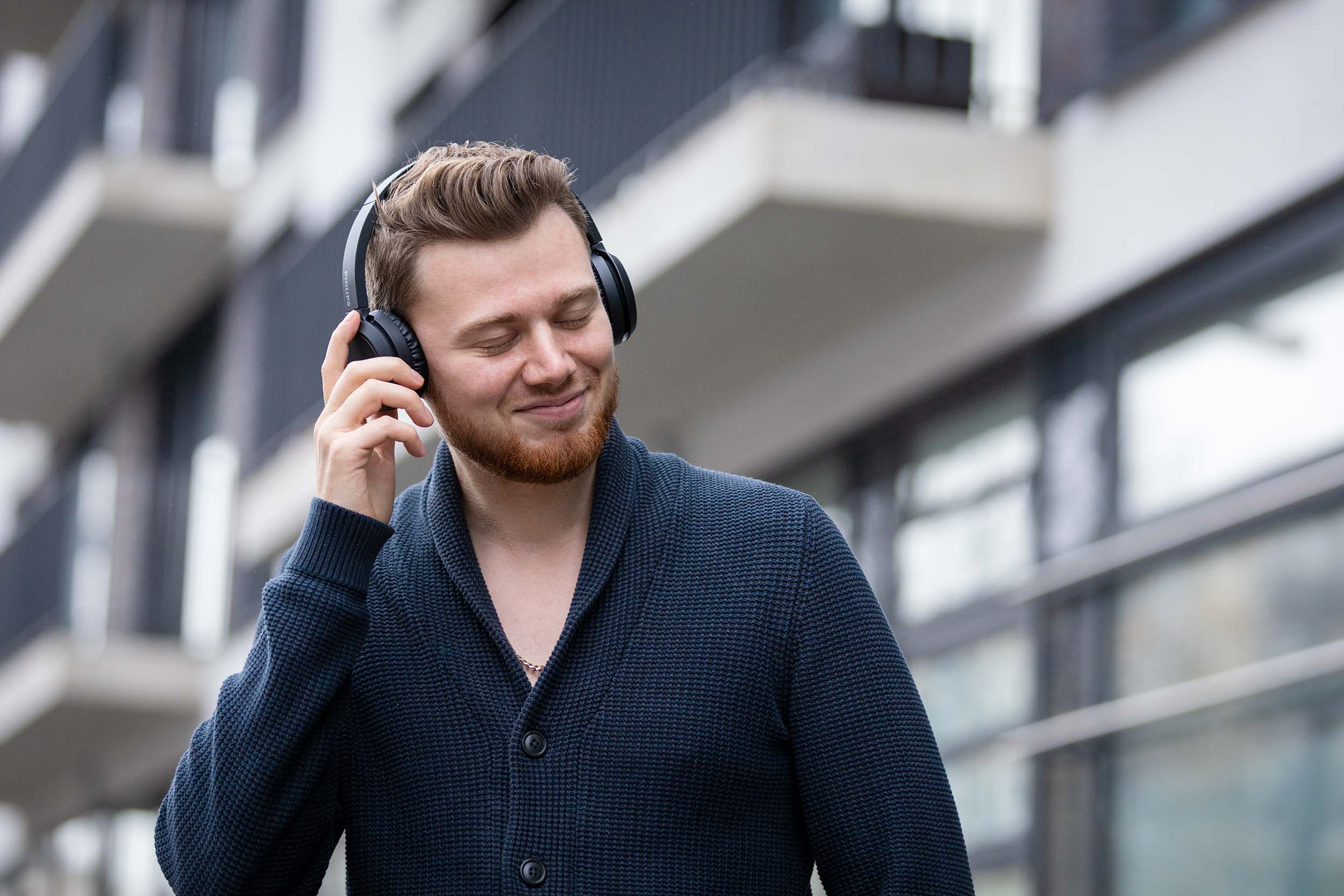 Dating-Foto eines jungen Mannes mit geschlossenen Augen, der vor einem städtischen, modernen Hintergrund glücklich lächelnd an seinen Kopfhörer greift.