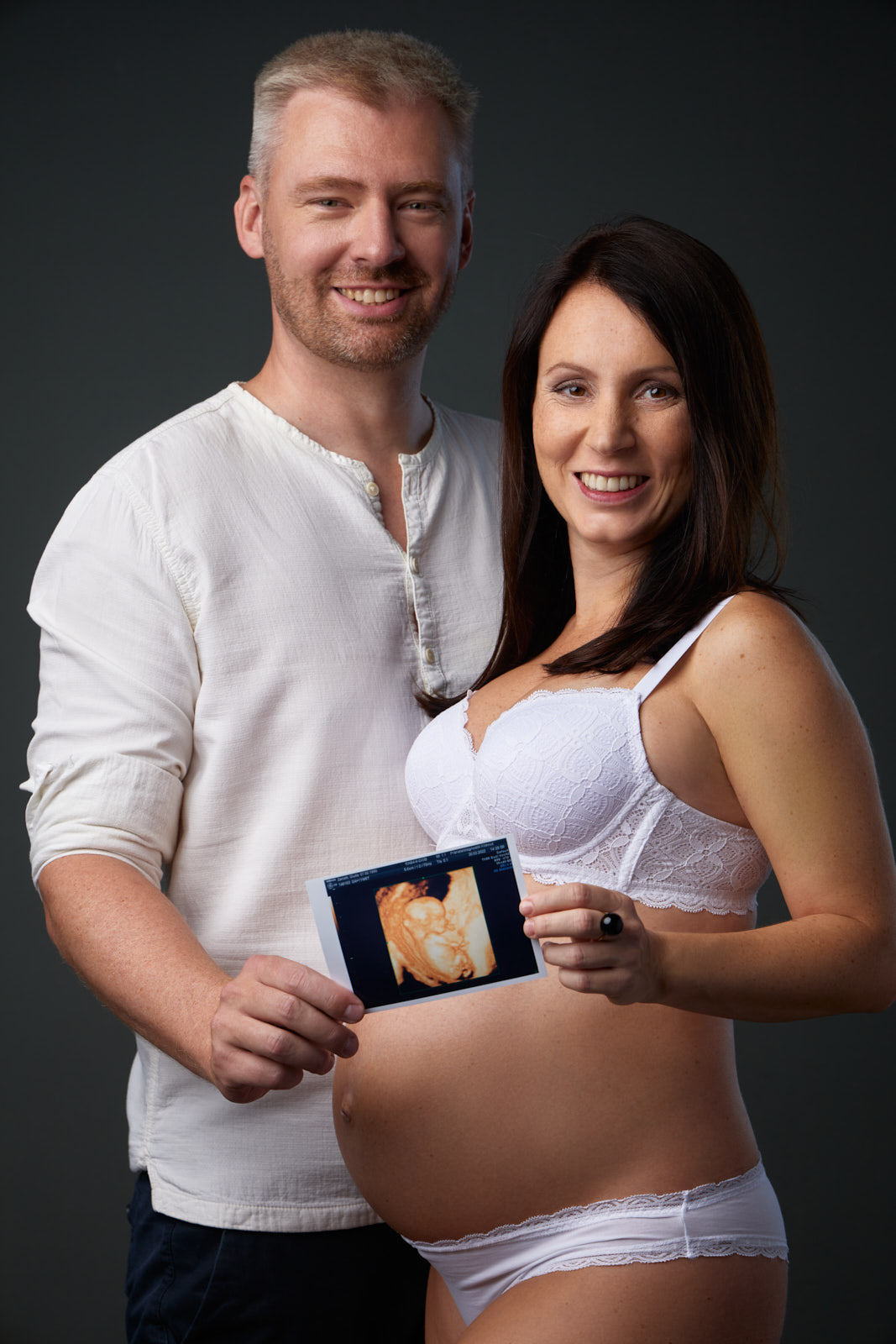 Ein freundlich lächelndes werdendes Elternpaar hält das Ultraschallfoto ihres Babys vor den dicken Babybauch in die Kamera. Die Mame trägt weiße Unterwäsche und der werdende Papa eine weißes kragenloses Leinenhemd.