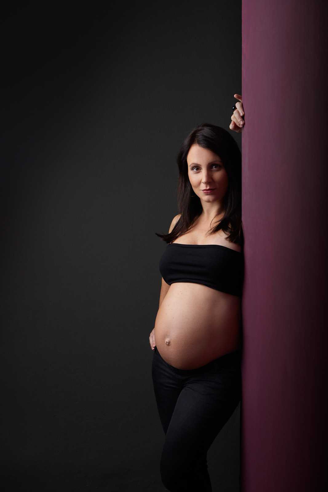Babybauchfoto einer Frau mit schwarzen langen Haaren in einer schwarzen Hose und einem schwarzen Bandeau lehnt heruasvordernd an einer lilanen Wand und Blickt souverän in die Kamera