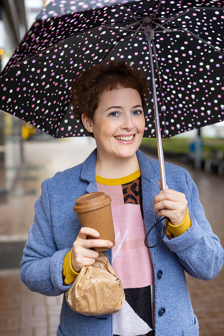 Dating-Foto einer jungen Frau mit kurzen lockigen Haaren die einen hellblauen Mantel trägt. Sie kommt gerade vom Bäcker mit Brötchen und einem Kaffeebecher in der Hand und trägt einen Regenschirm.
