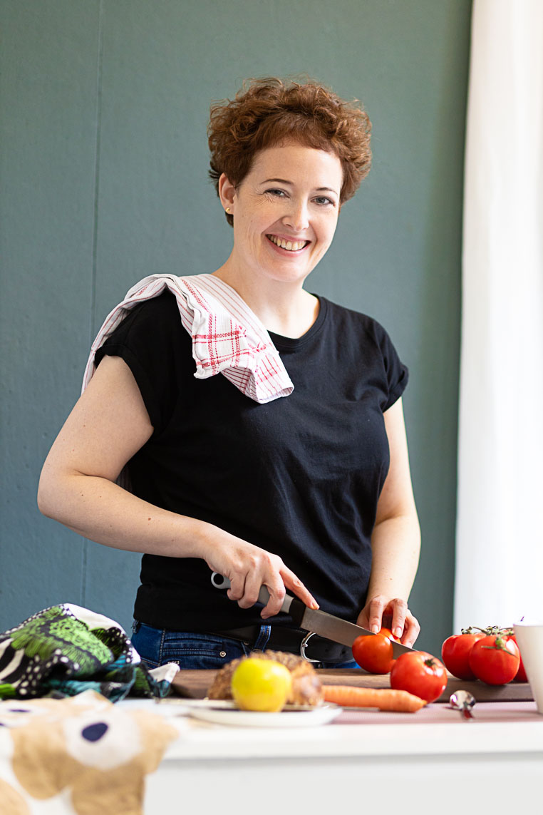 Dating-Foto einer junge, lächelnden Frau mit kurzen lockigen Haaren, die gerne Kocht beim Gemüseschniden in der Küche. Sie trägt ein schwarzes T-Shirt und steht vor einer grün-blauen Wand.