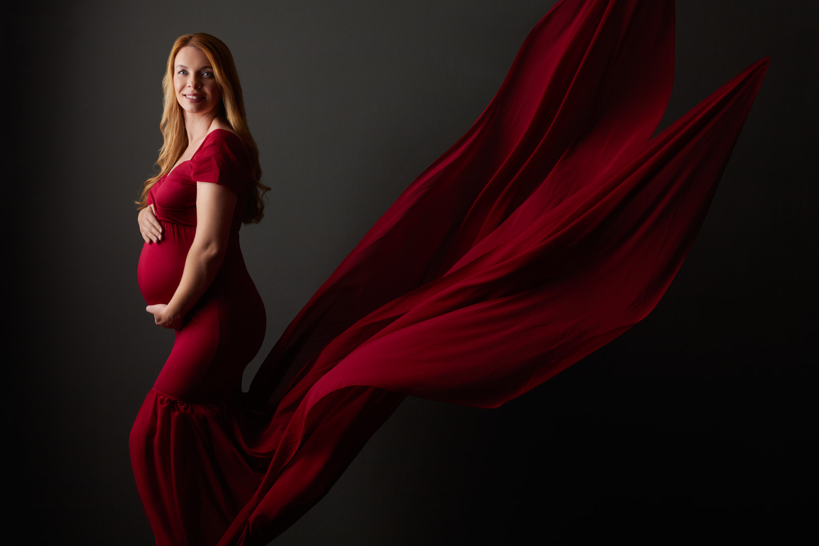 Fotoshooting einer schwangeren, glücklich lachenden, blonden Frau in einem lange wehenden rotem Kleid