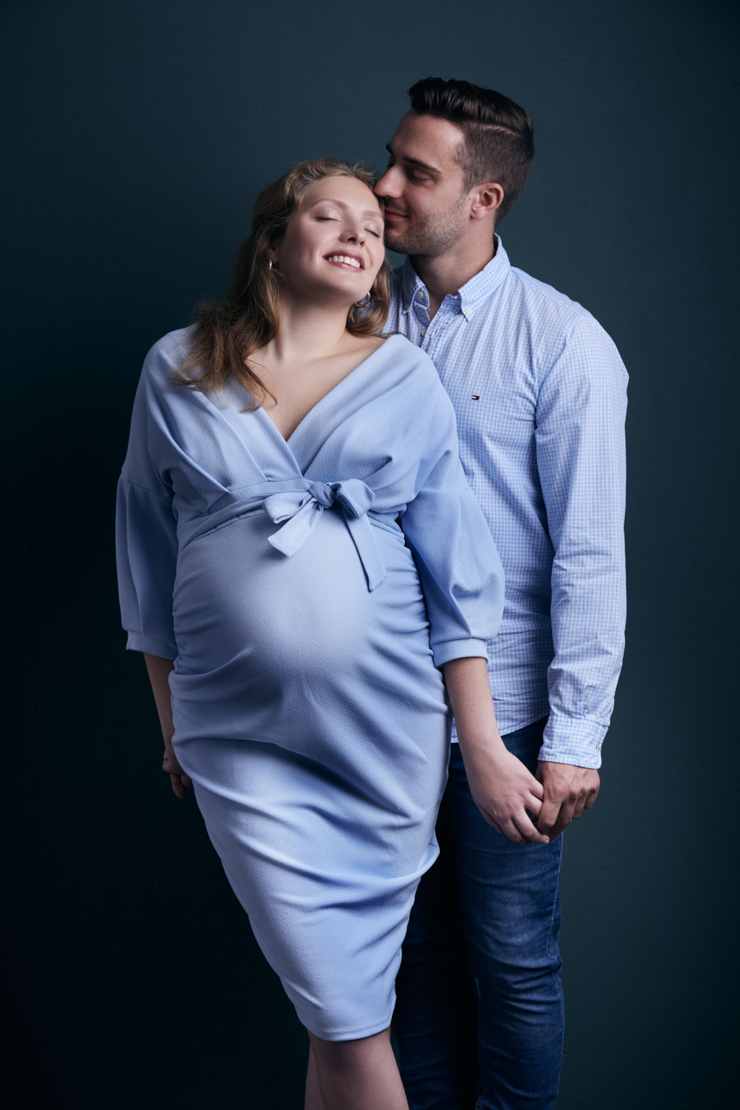 Fotoshooting eines werdenden Elternpaares. Die schwangere, glücklich, lächelnde junge Frau wird von Ihrem Mann auf die Schläfe geküsst. Sie trägt ein elegantes hellblaues Kleid und er ein passendes Hemd.