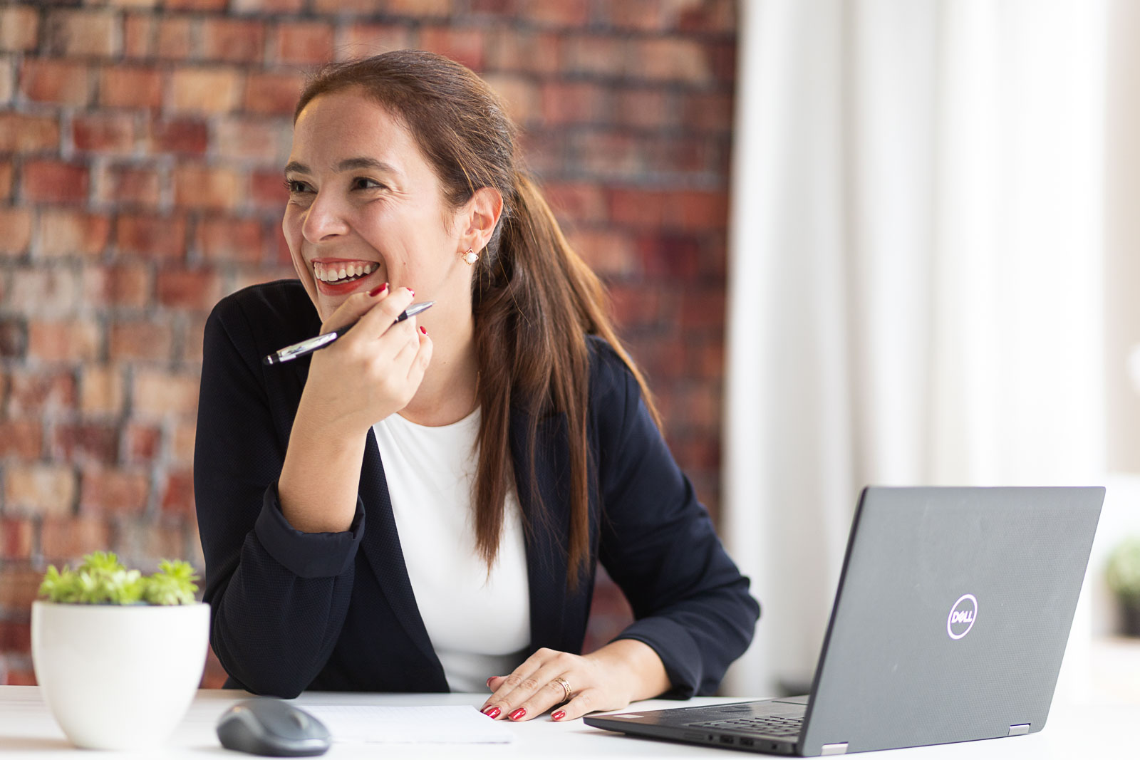 Dating-Foto einer junge, lachenden Frau mit lange, braunen Haaren zu einem Pferdeschwanz zusammengebunden. Sie sitzt mit einem Stift in der Hand in ihrem Büro an einem Schreibtisch mit Laptop und lacht freudig. Im Hintergrund ist eine rohe Backsteinwand.