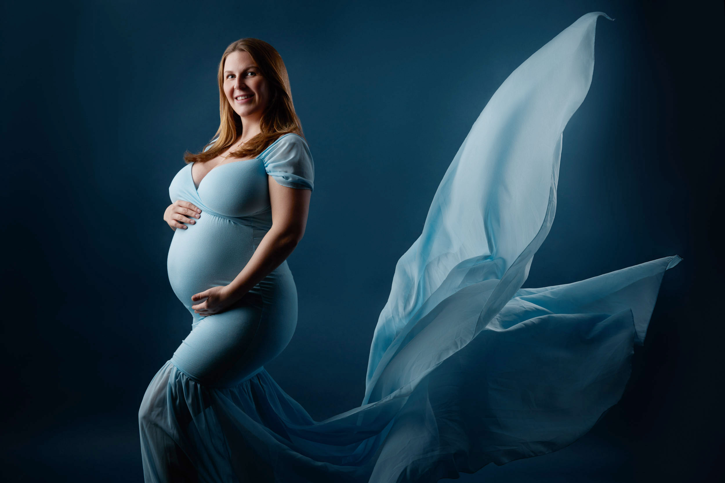Fotoshooting einer schwangeren, glücklich lachenden,blonden Plus-Size Frau in einem bleune Kleid, das elegant wie ein Meerjungfauenschwanz weht.