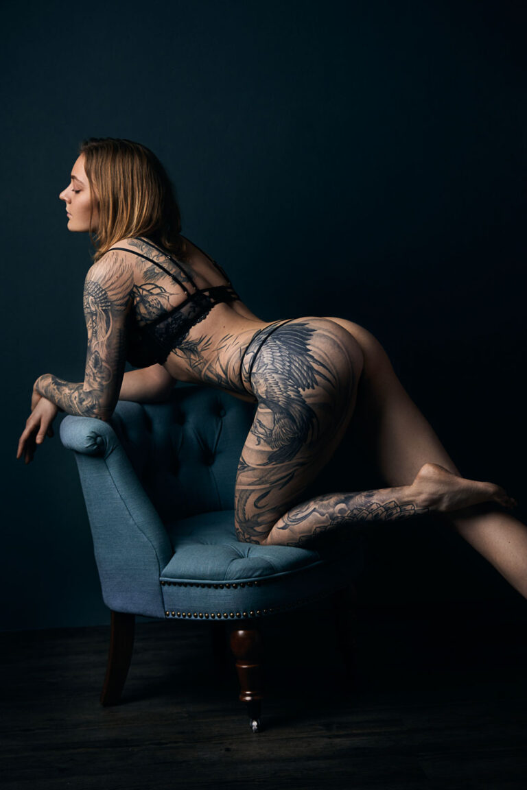 Akt-Fotoshooting einer braunhaarigen tatowierten jungen Frau die in schwarzer Unterwäsche vor einer petrolfarbenen Wand auf einem ebenfalls blauen Sessel elegant kniet.