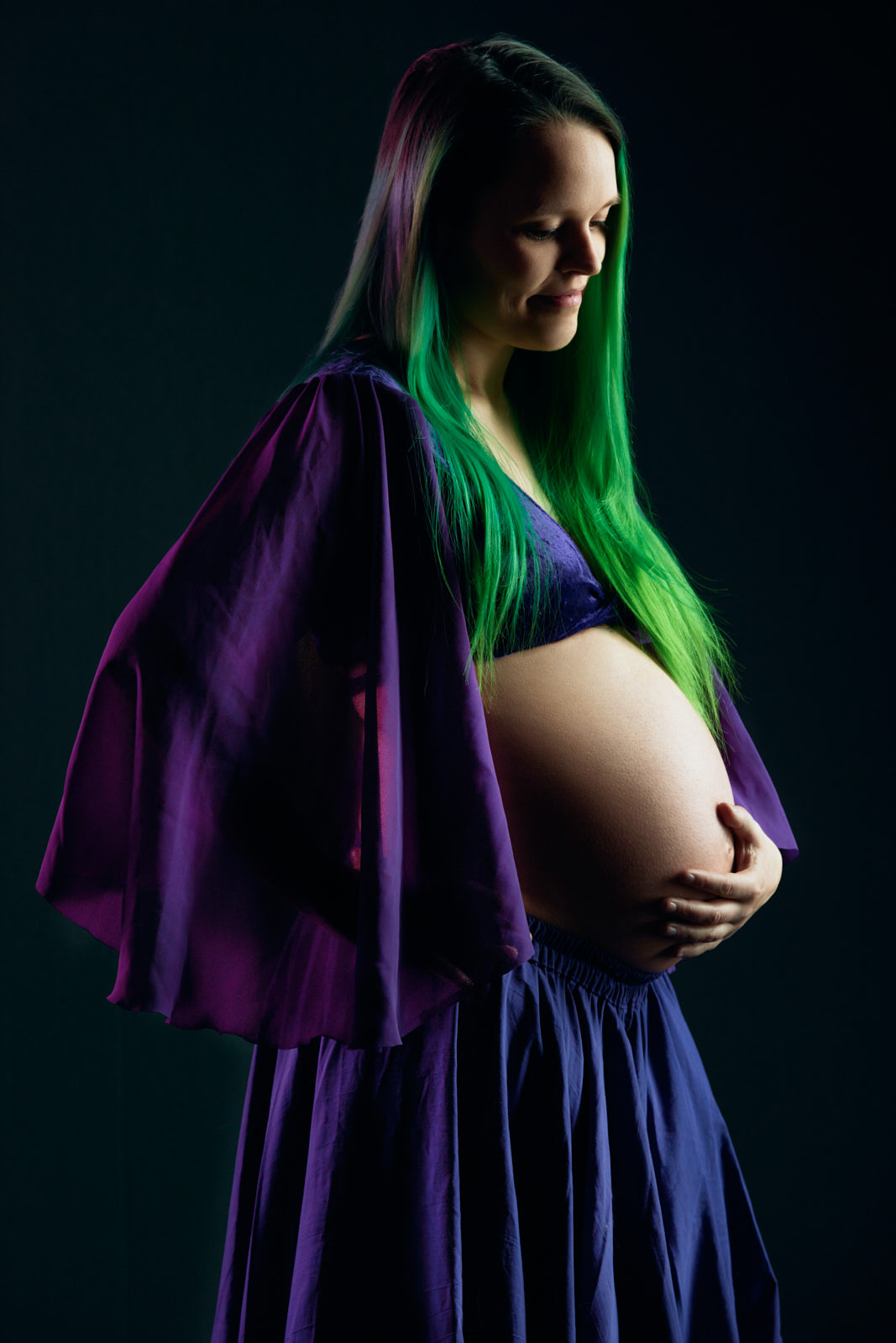 Babybauchfoto einer schwangeren Frau mit grün gefärbten, langen Haaren in einem blauen Rock, die freudig, sinnend auf ihren Babybauch schaut.