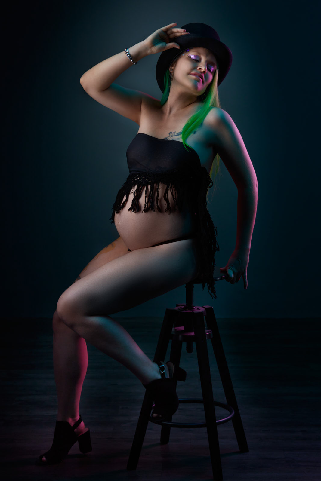 Babybauchfoto einer schwangeren Frau mit grün gefärbten, langen Haaren die auf einem hohen Hocker sitzt, sich zurücklehnt und den Zylinder hält, den sie auf dem Kopf trägt.