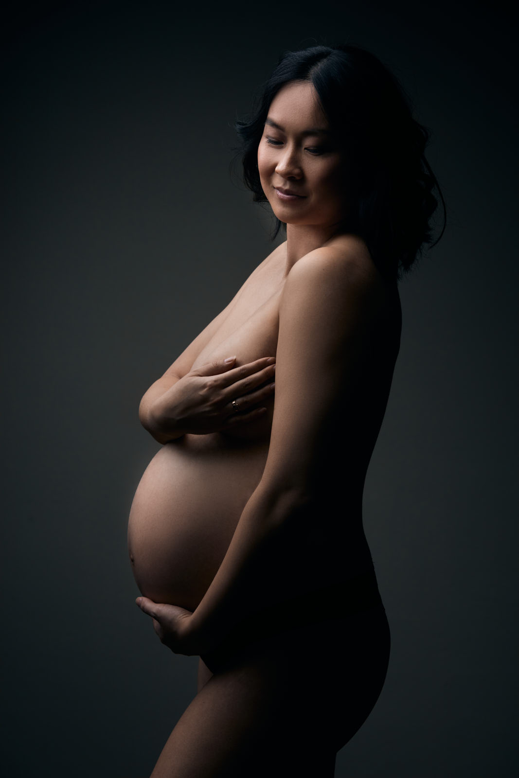 Babybauchfoto einer asiatisch aussehnden Frau mit schwarzen Haaren, die nackt seitlich steht und Ihre Brüste und Bauch mit den Händen bedeckt