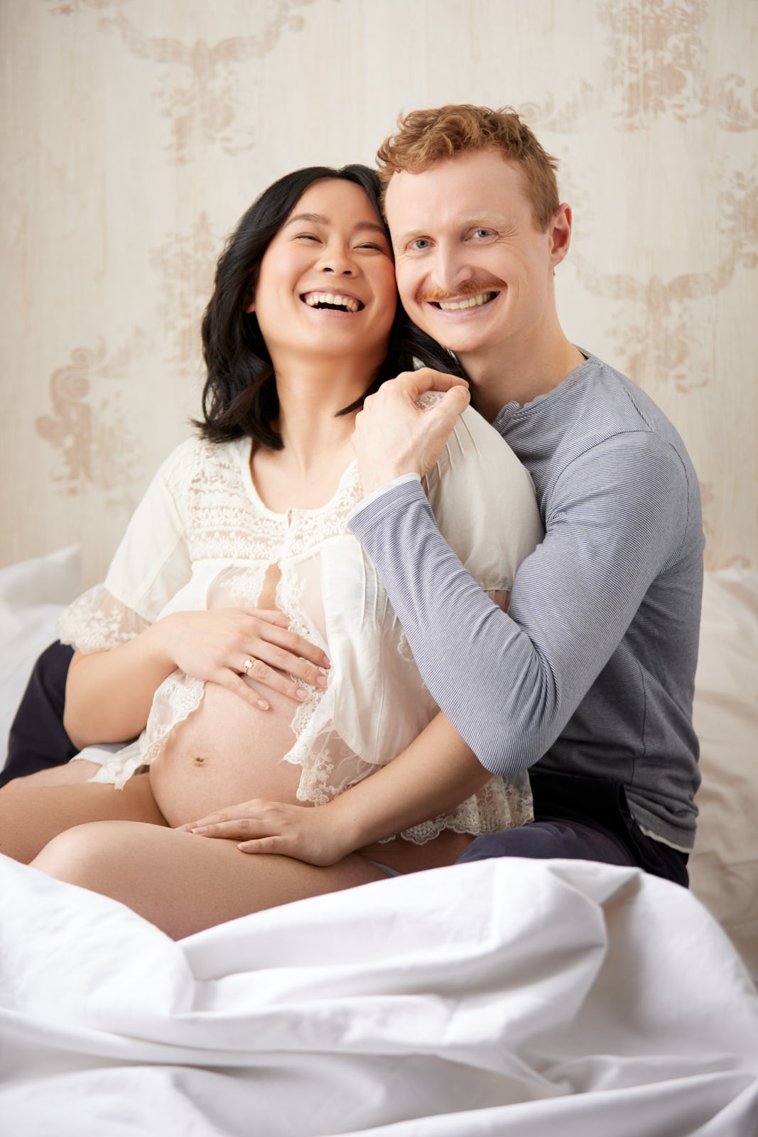 Babybauchfoto einer asiatisch aussehnden Frau die zusammen mit ihrem rothaarigen Mann vor einer hellen Wand seitlich auf dem Bett sitzen. Sie lehnt sich glücklich lachend an ihn an, der in die Kamera schaut und sie an der Schulter hält.
