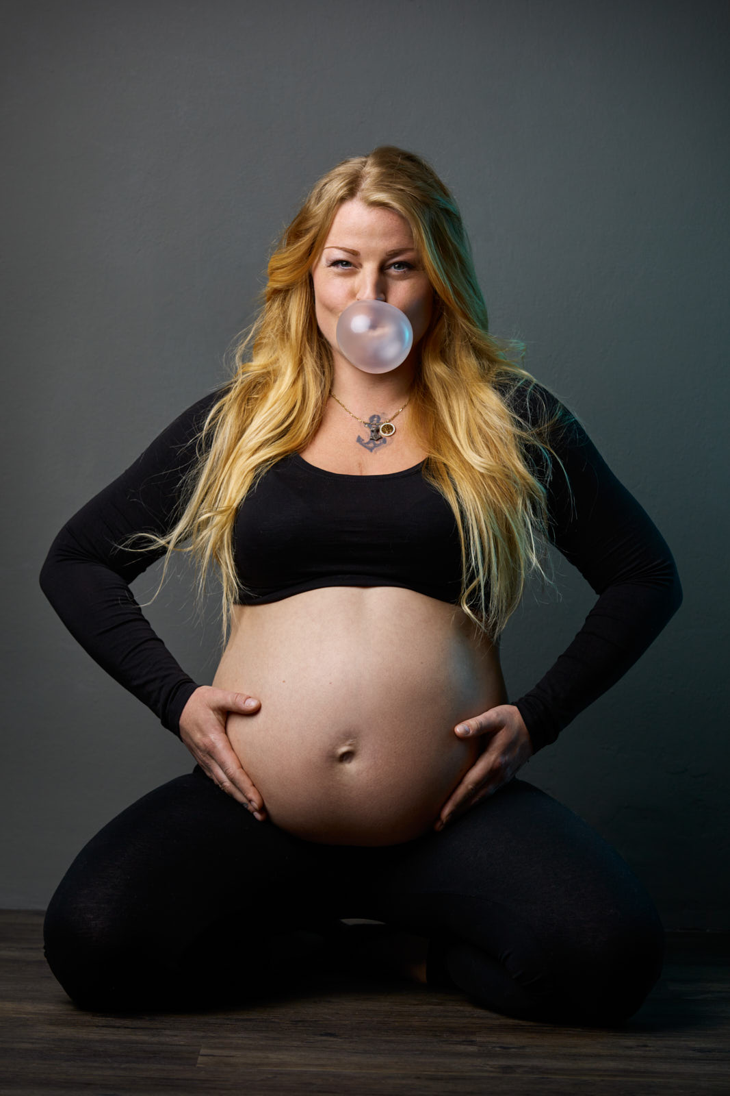 Babybauchshooting einer schwangeren, werdender Mutter, die in einem Gymnastikoutfit, vor einer graublauen Wand auf dem Boden sitzt und lachend eine Kaugummiblase macht und Ihren Bauch hält.