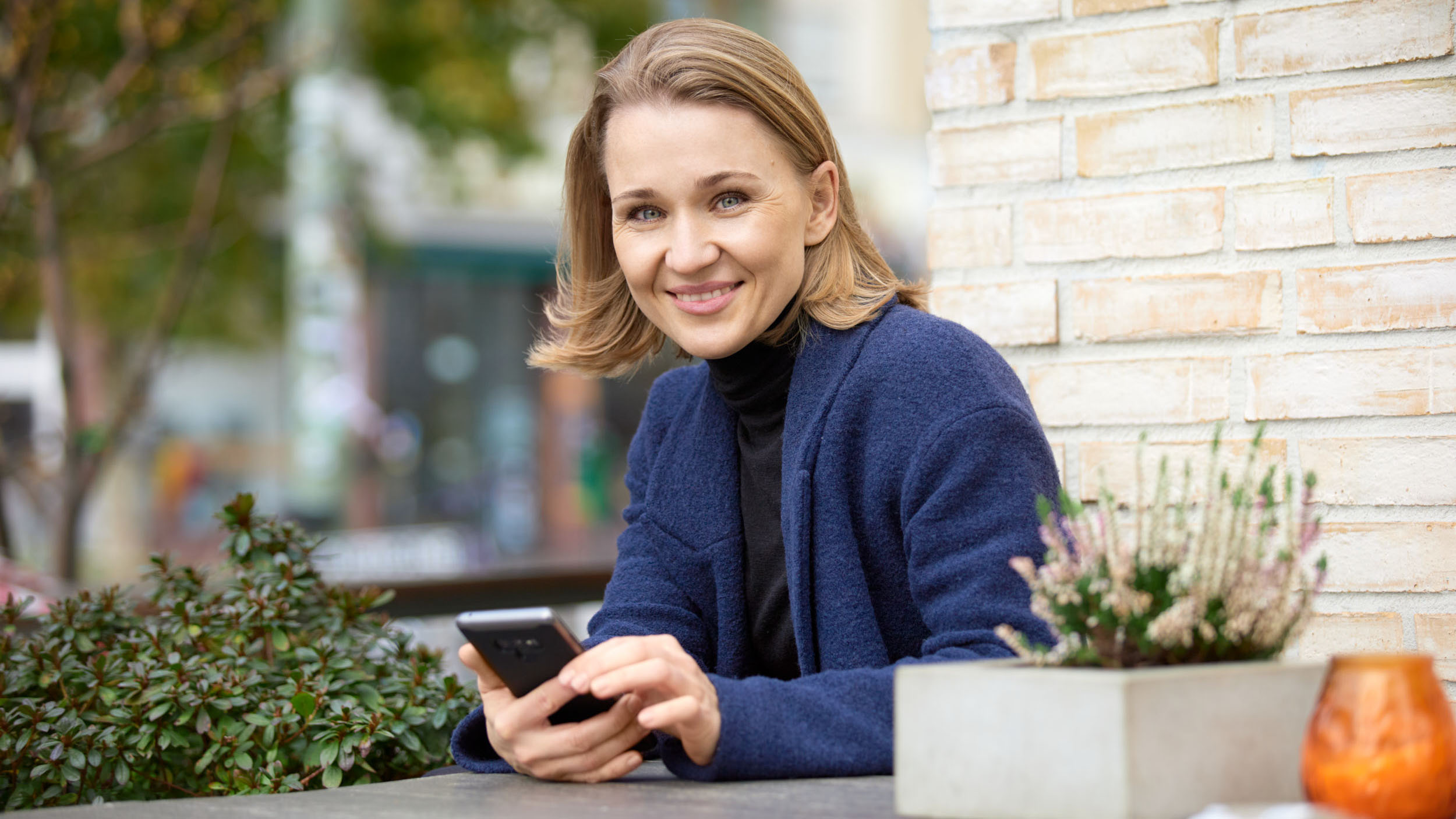 Dating-Foto einer blonden jungen Frau mit schulterlangen Haaren, die mit ihrem Handy in der Hand am Tisch eines Straßenkaffees sitz