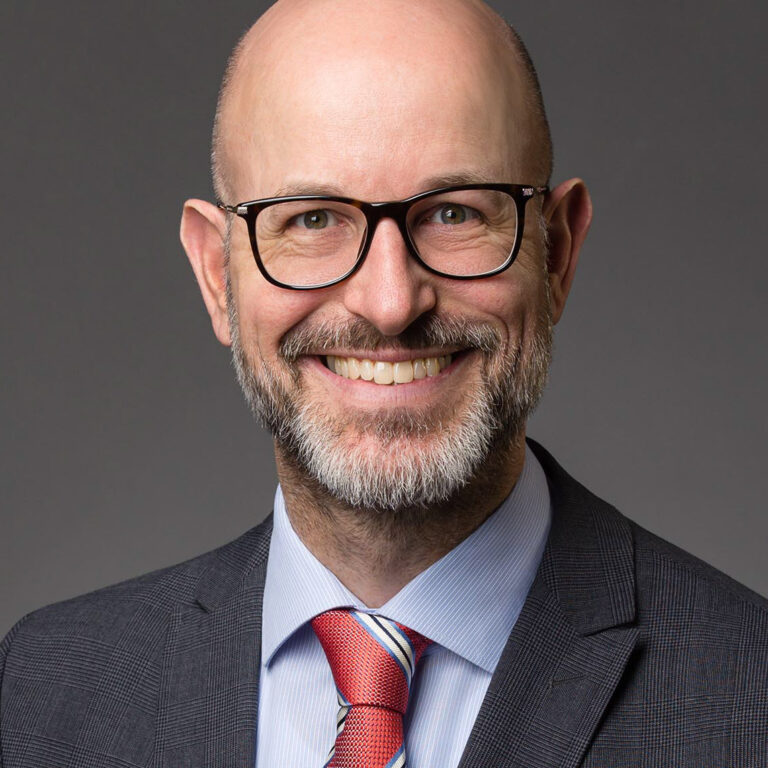 Modernes Profilfoto für Xing und LinkedIn eines freundlich lächelnden Mannes mit Brille, Vollbart und roter Krawatte vor grauem Hintergrund.