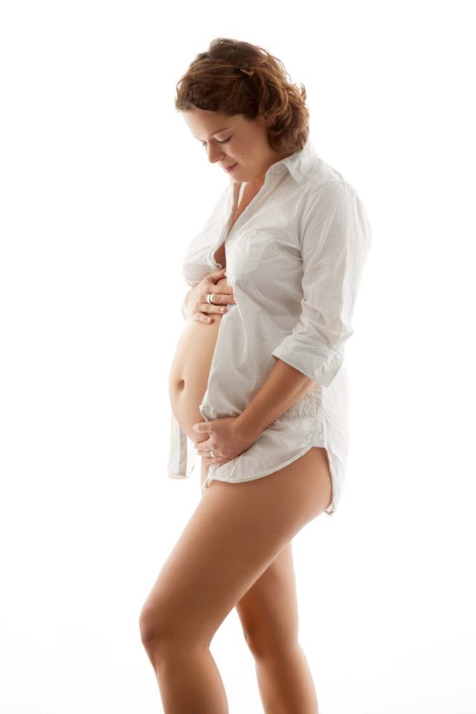 Schwangere Frau trägt ein weißes Männerhemd vor weißem Hintergrund und schaut auf Ihren Babybauch