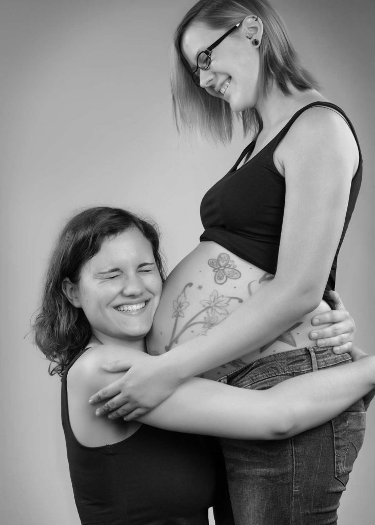 Foto eines lesbischen Paar. Eine Frau umarmt glücklich lächelnd den Babybauch ihrer schwangeren Freundin