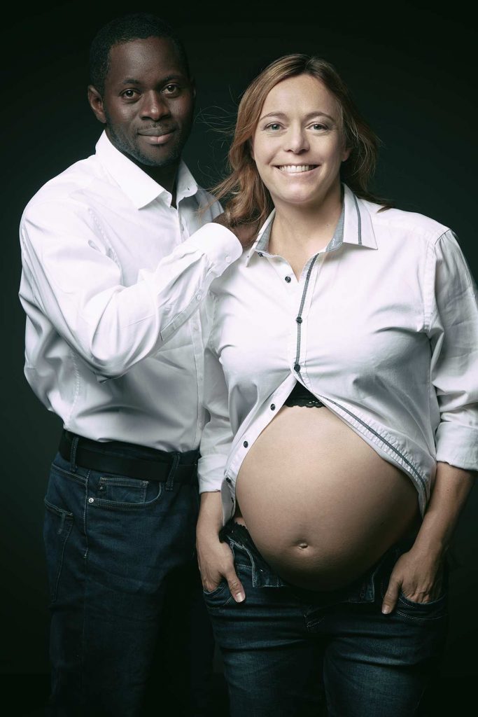 Dunkelhäutiger, werdender Vater und seine schwangere Frau mit Babybauch haben weiße Hemden an und blicken direkt in die Kamera
