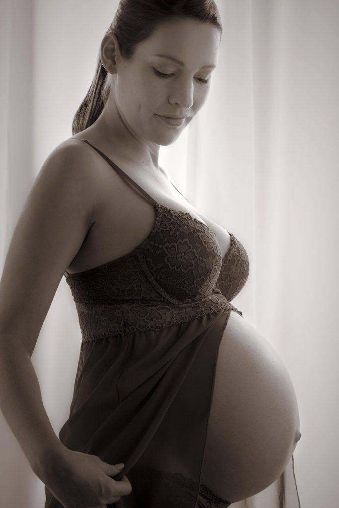 Schwarz-weiß Foto einer schwangeren Frau. Sie steht vor einem hellen Vorhang in einem Negligee und blickt auf Ihren Bauch