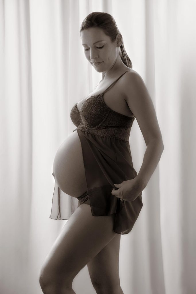 Schwarzweiß Foto einer schwangeren Frau in einem Negligee die vor einem Vorhang steht und auf Ihren Bauch herunter blickt