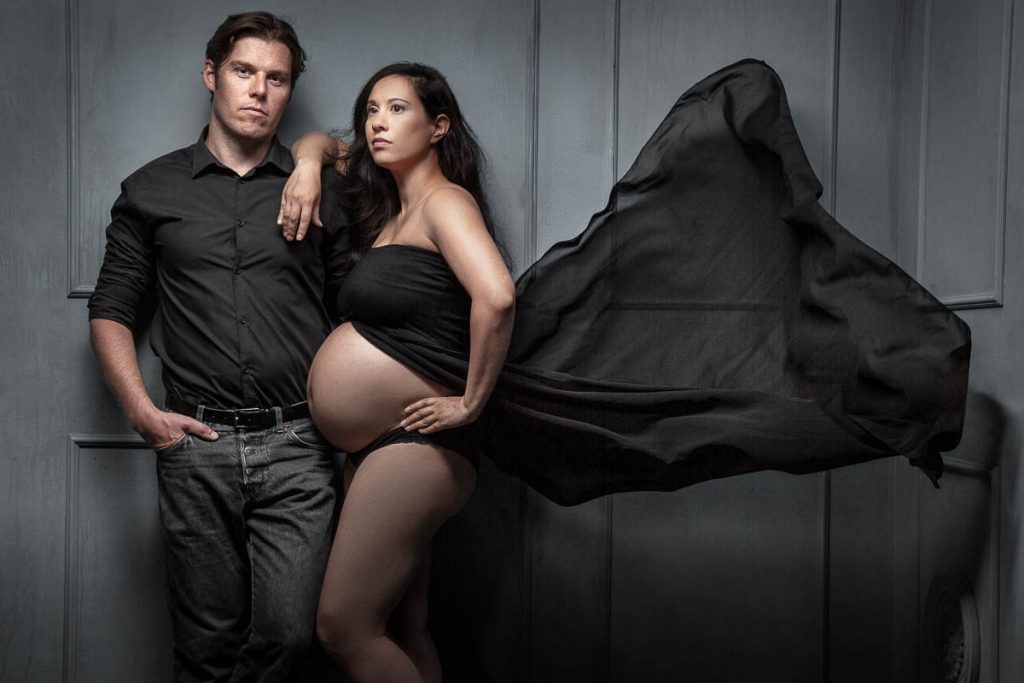 Cooles Babybauchfoto Paarfotoshooting einer schwangeren Frau zusammen mit dem werdenden Vater. Die Schangere stütz sich lässig auf die Schulter ihres Mannes, währen ein schwarzes Tuch hinter weht.