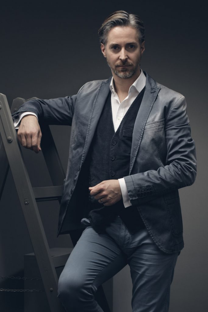 Künstlerportrait eines Schauspielers, der ein Drycorn Jacket trägt und sich auf eine graue Leiter stützt